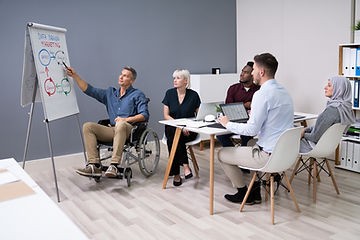 A man who is using a wheelchair is leading a meeting and pointing at a flip chart. 4 people are seated around a conference table listening attentively.