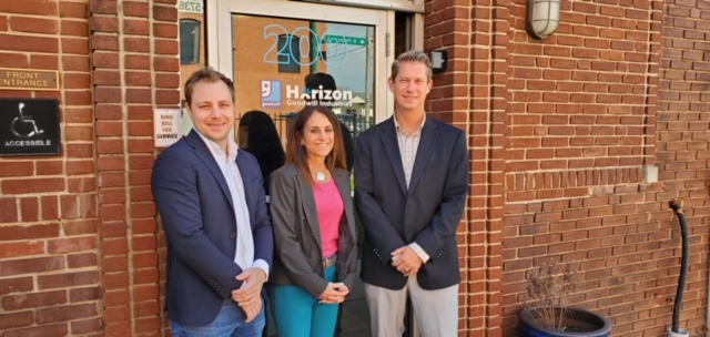 Three people standing in front of a brick building entrance with a sign that reads ‘Horizon Goodwill Industries.’ They are dressed in business attire, smiling at the camera.
