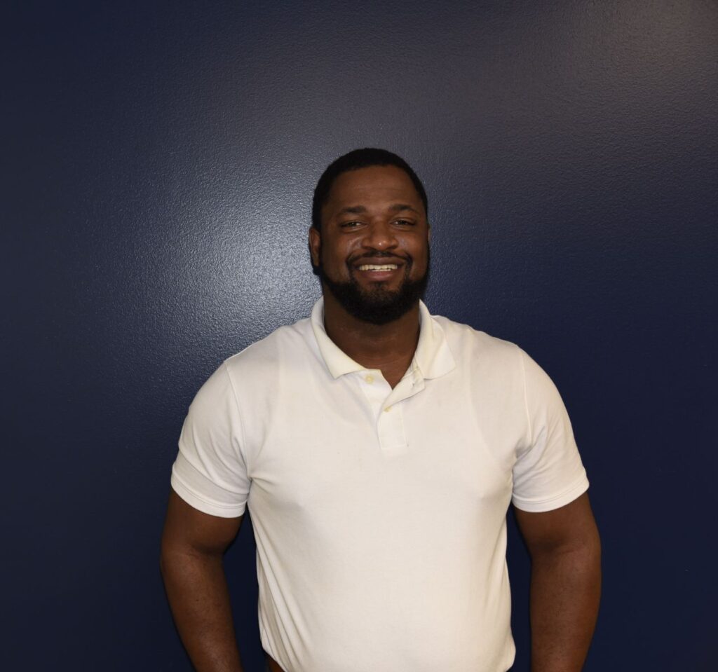 Man smiling for a portrait in front of a dark blue wall, wearing a short-sleeved white polo shirt.