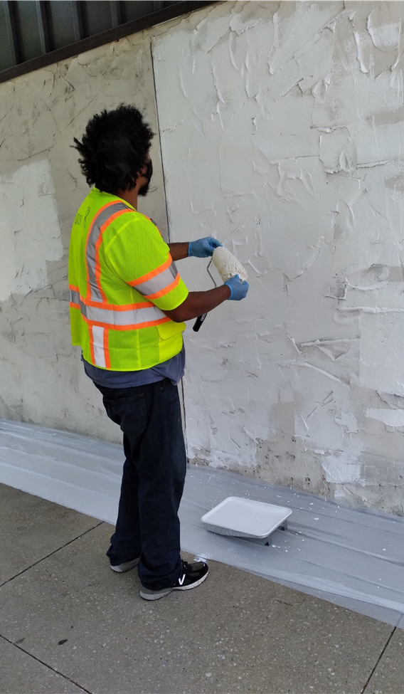 A worker wearing a bright yellow safety vest, gloves, and jeans is painting a textured wall with a roller. A paint tray with white paint is on the ground nearby.