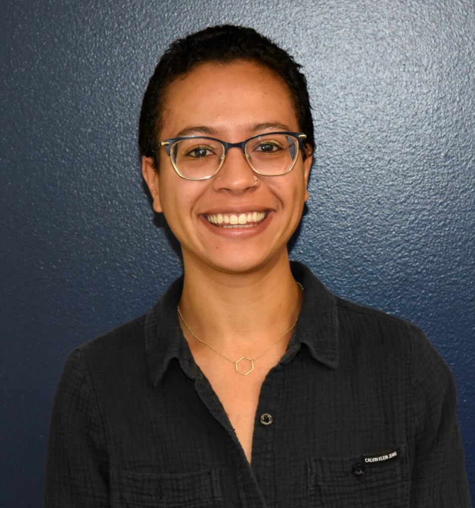 Woman smiling for a portrait in front of a dark blue wall, wearing glasses, a nose ring, a dark button-up shirt, and a geometric necklace.