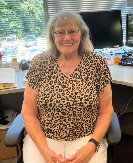 Woman smiling while seated at an office desk, wearing glasses and a leopard print shirt with white pants. A computer, office supplies, and a window with sunlight and trees outside are in the background.