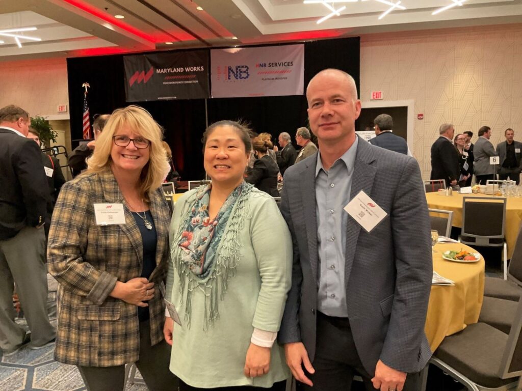 Three people smiling and posing together at a formal event with round tables and yellow tablecloths. They are wearing business attire with name badges. In the background, other attendees are mingling, and banners for Maryland Works and sponsors hang on the stage.
