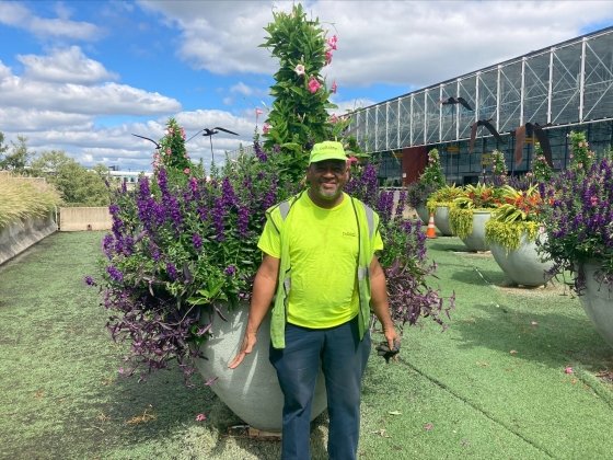 Mikey, in a flourescent yellow-green shirt and safety jacket, standing outside next to large flower bushes, as part of the Employment Works Program.