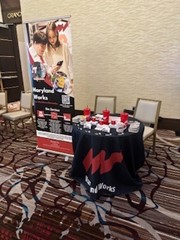 An event display table covered with a black Maryland Works tablecloth featuring the red ‘MW’ logo. The table is set with promotional items including red cups and brochures. A tall banner with images of people and text about Maryland Works stands beside the table in a carpeted conference space with chairs in the background.