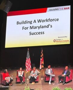 A panel of five speakers seated on stage in front of a large screen displaying the words: ‘RAISING THE BAR. Building A Workforce For Maryland’s Success.’ The event is sponsored by the Center for Workforce Inclusion, with the organization’s logo visible on the screen. The U.S. and Maryland state flags stand on stage between the panelists.