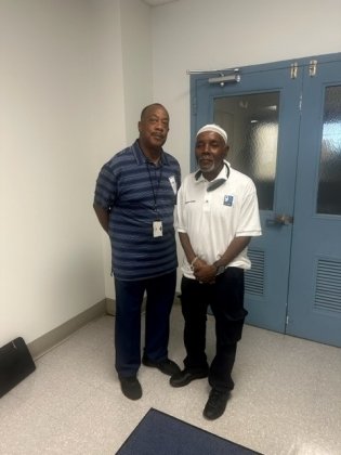 Two men standing indoors and smiling at the camera. One is wearing a blue striped shirt and dark pants, and the other is wearing a white collared shirt with a logo, dark pants, and a white cap. They are standing in front of blue double doors.