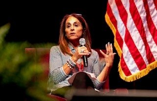 A Maryland Works, Inc. representative seated on stage speaking into a microphone during a panel discussion. She is gesturing with her hand while addressing the audience. The U.S. flag is visible beside her.