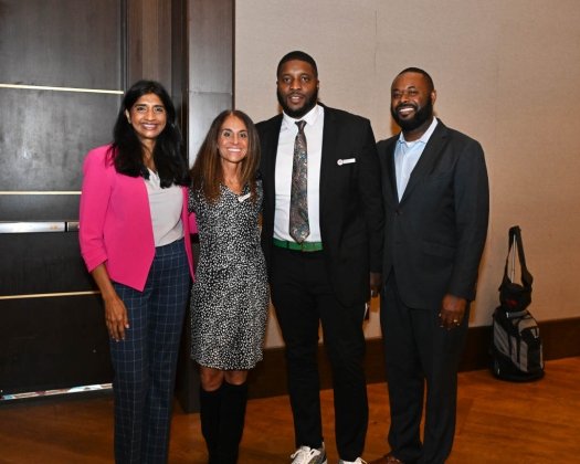 Four adults stand together indoors, smiling at the camera. From left to right, a woman in a pink blazer and plaid pants, a woman in a patterned dress with knee-high boots, a man in a dark suit with a patterned tie, and a man in a dark suit with an open-collar shirt. They appear to be at a professional or social event.