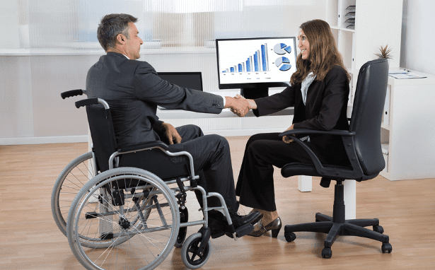 A man in a wheelchair and a woman seated in an office chair shake hands across a desk. Both are dressed in business attire, and a computer screen in the background displays charts and graphs, suggesting a professional meeting or partnership.