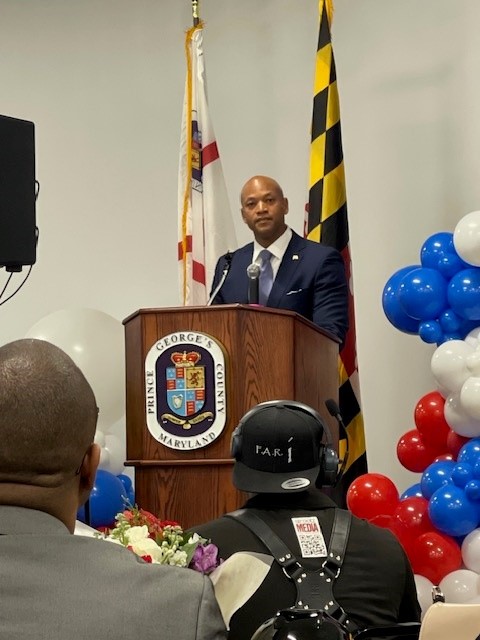 Maryland Governor speaking at a podium with the Prince George’s County, Maryland seal. The U.S. flag and Maryland state flag are displayed behind him, and red, white, and blue balloon decorations frame the scene as audience members listen.