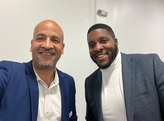 Two men smiling for a photo indoors against a plain white wall. Both are wearing blazers; the man on the left is in a blue jacket with a white shirt, and the man on the right is in a plaid jacket with a white turtleneck.