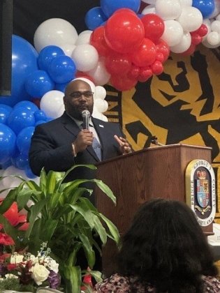 A man in a suit speaks at a podium with the Prince George’s County, Maryland seal during a ribbon cutting ceremony. Red, white, and blue balloons decorate the background along with a black and gold banner. Audience members are seated in front, and plants and flowers are arranged near the stage.
