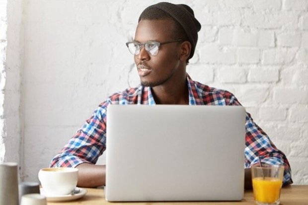African American man wearing a black knit hat, glasses, and a flannel work shirt, looking out the window and writing on his laptop.