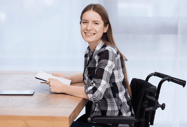 woman sitting in a wheelchair at a desk smiles to the camera, taking a break from reading her note pad
