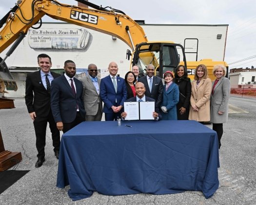 Governor Wes Moore sits at a table outdoors holding a signed document, surrounded by a group of smiling officials and colleagues. Behind them is a large yellow JCB excavator and a building with a mural that reads ‘The Frederick News-Post.’