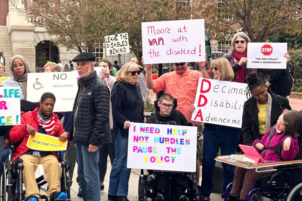 A group of unhappy people peacefully protesting and holding signs such as "Moore at war with the disabled!", "I need help not hurdles pause the policy", "STOP the DDA Policy; form a workgroup!".