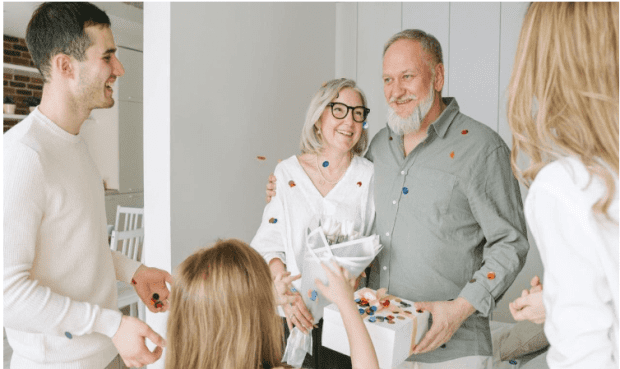 A smiling older couple, with the man holding a gift box, is warmly greeted by family members in a bright home. The group is tossing colorful confetti, creating a celebratory atmosphere.